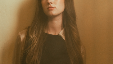 Promotional photo for "Freshman Year" which sees Isabel Dumaa standing against a beige wall, looking slightly to the left, with her brown hair cascading down past her shoulders and in front of her black dress. Her hands are behind her back.