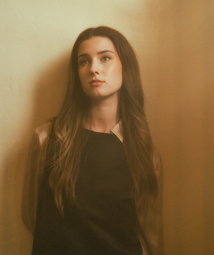 Promotional photo for "Freshman Year" which sees Isabel Dumaa standing against a beige wall, looking slightly to the left, with her brown hair cascading down past her shoulders and in front of her black dress. Her hands are behind her back.