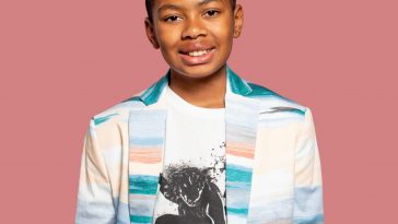 Phoenix Nicholson at 11 years old standing in front of a pink background, wearing a white shirt with a black print of a skater on it, underneath a smart jacket that is white with watercolour paint horizontal lines of blue, pink, green, and beige on it. He is smiling at the camera.