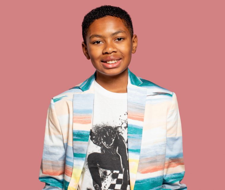 Phoenix Nicholson at 11 years old standing in front of a pink background, wearing a white shirt with a black print of a skater on it, underneath a smart jacket that is white with watercolour paint horizontal lines of blue, pink, green, and beige on it. He is smiling at the camera.