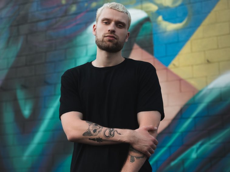 Promotional photo for "I Got Lost In Berlin" which sees Sikdope with short bleached blonde hair and a natural-brown beard, wearing a black t-shirt, standing in front of an art memorial wall. He is holding his left arm with his right, gripped just below the elbow, with his right arm coming across his body, showcasing his tattoo art on his forearm.