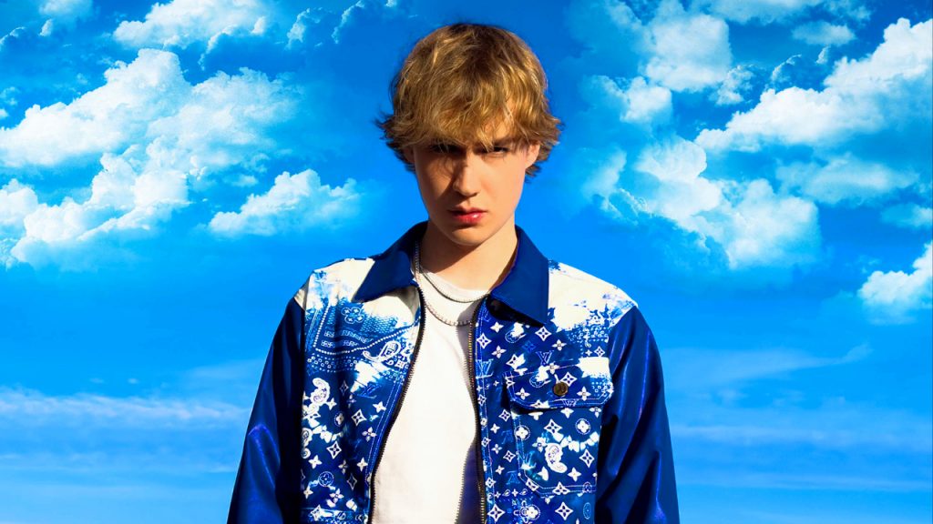 Promotional photo for "Sky High" which sees Alex Shade in front of a blue sky with light blue clouds in the background. His dirty blonde hair is a messy mid-short length and he is wearing a white t-shirt under a dark blue jacket.