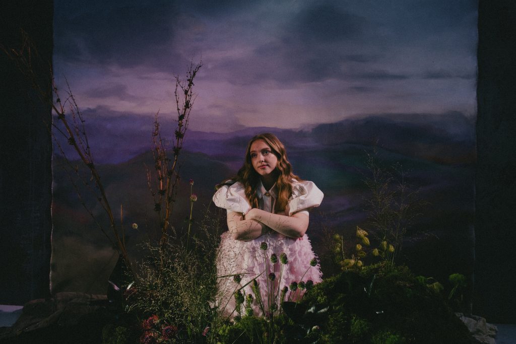 Promotional image for "Greek Street" which sees Alice Auer on a set posing for the camera in a white dress behind some grass, while there's a background of a road with a purple sky and fields on either side of her.