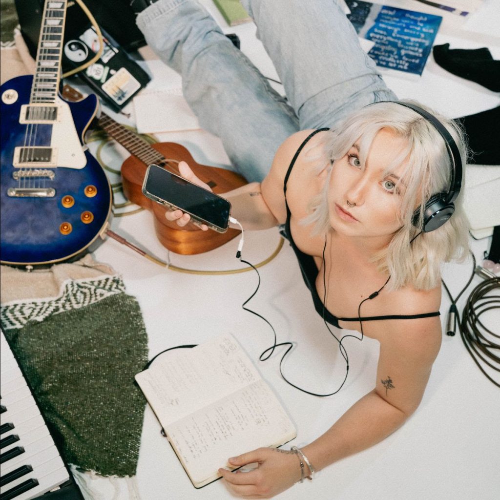 Promotional photo for "comfortable - voice memo" which sees Ashlynn Malia lying down on a white blanket on a green carpet looking up at the camera as she has black headphones on, a black strap top paired with light blue jeans. She has her songwriting pad in front of her, along with a keyboard, as well as a blue guitar and a brown guitar to the top-left of the image, as well as recording equipment spread around her.