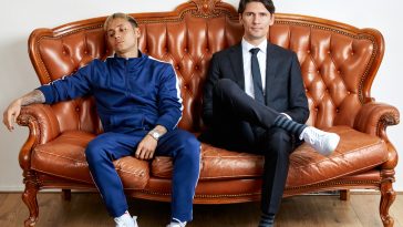 Promotional photo for "Les Pays Bass" which sees Bassjackers sitting on a brown leather couch with a white-walled room and wood flooring with a red carpet rug. Marlon Flohr is on the left wearing a blue tracksuit with white sneakers, and Ralph van Hilst is on the right wearing a black two-piece suit with a navy tie and white sneakers.