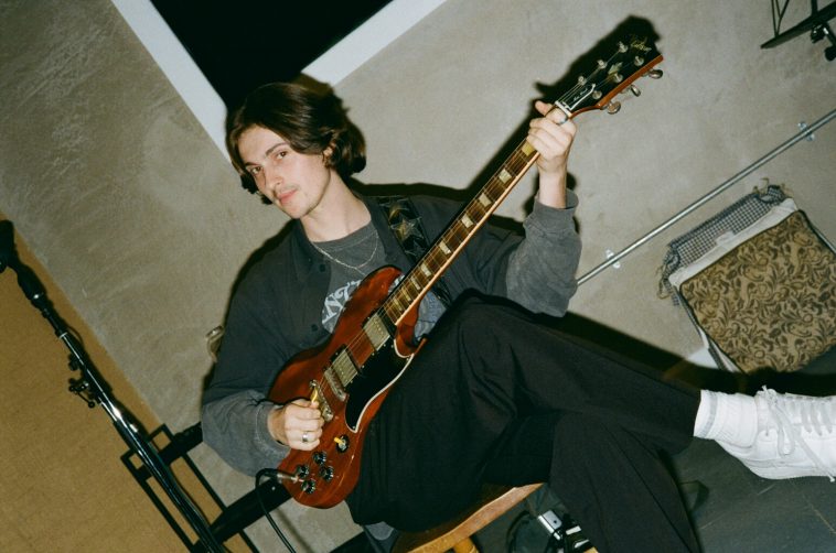 Promotional photo for "Blood" which sees bryden, who has just-under shoulder-length dark brown hair, sitting on a chair with his guitar across his lap with his hands on it as if he's playing it. His head is turned to the left and he's slightly smiling. He's wearing a sea-green shirt over a grey t-shirt and some black cargo trousers.