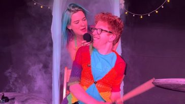 Promotional photo for "Honeymoon Love" which sees MuMu behind a ginger-haired man who is playing the drums. She's looking at him over his shoulder as he looks at her through his wide-brimmed glasses as he grins with happiness. He is wearing an orange cardigan over a sky-blue shirt, with drumsticks in his hands. MuMu has blue hair down past her shoulder and is backed by a purple background.