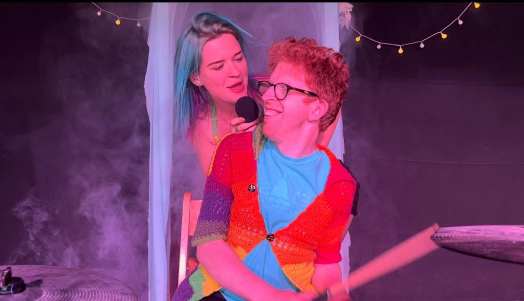 Promotional photo for "Honeymoon Love" which sees MuMu behind a ginger-haired man who is playing the drums. She's looking at him over his shoulder as he looks at her through his wide-brimmed glasses as he grins with happiness. He is wearing an orange cardigan over a sky-blue shirt, with drumsticks in his hands. MuMu has blue hair down past her shoulder and is backed by a purple background.