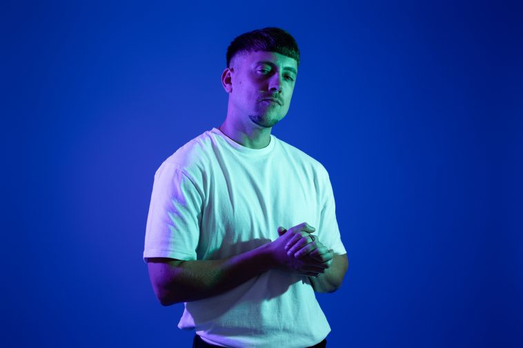 Promotional photo for "Find the Time" which sees Axel Boy standing in front of a blue background, he is wearing a white t-shirt and facing slightly to the right. He has his hands clasped together in front of him.