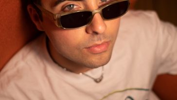 Promotional photo for "Quarter Century" which sees Charles On TV wearing an off-white t-shirt with a black logo of Arco Lounge displayed on the front, paired with a pair of shades and a silver necklace. His short curly black hair is slicked somewhat back and he seems to be sitting on a wooden seat against a dark orange-brown wall.