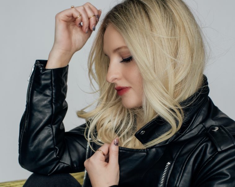 Press photo of Claire Guerreso who just dropped her new single "The Let Down". She is wearing a leather biker jacket and is facing to the left with her head slightly down as he blonde hair falls past her arms. Her right arm is bent at the elbow with her hand close to moving her hair out of her face, while the left hand is gripped onto her jacket.