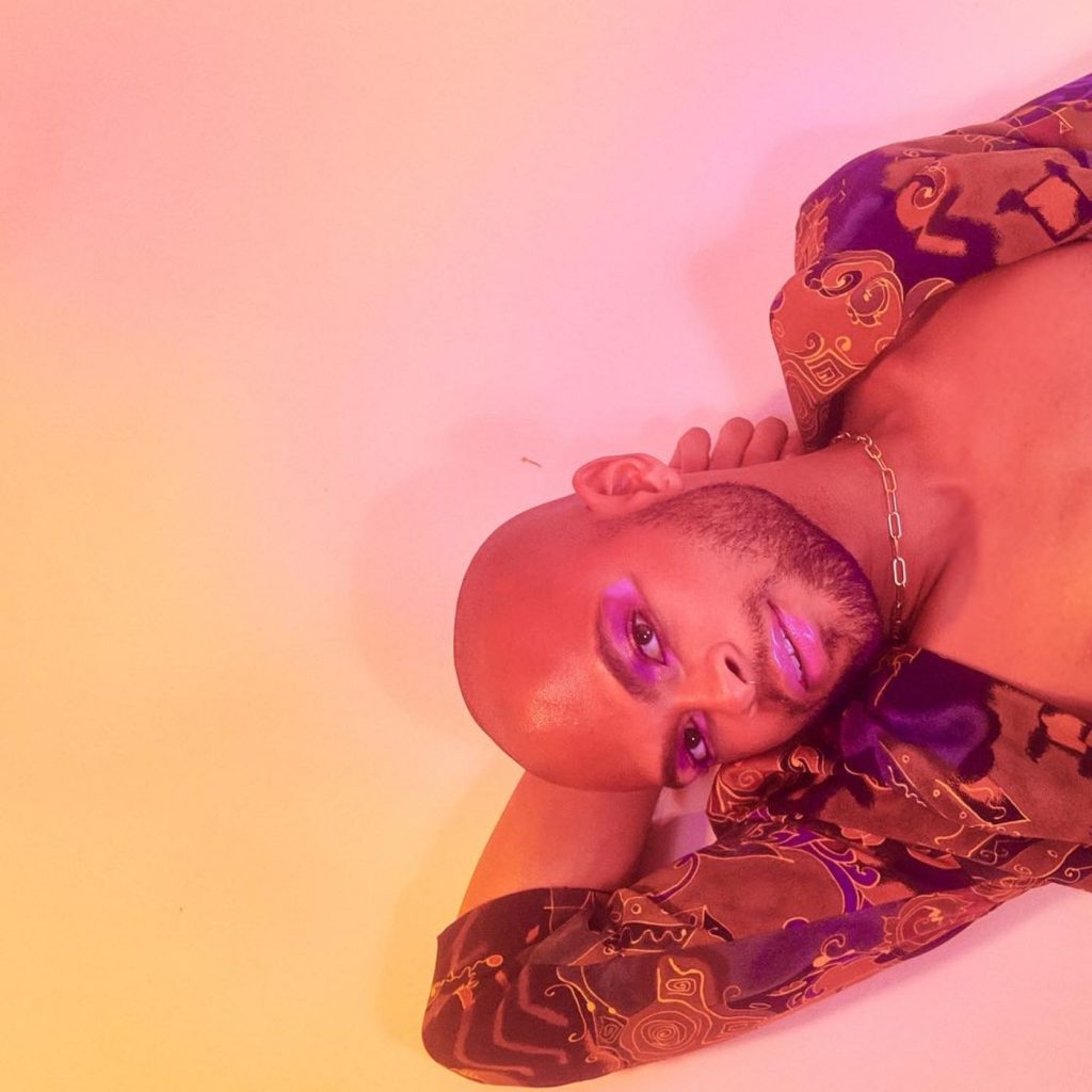 Single cover artwork for "romantic fury" which sees nasir mf. lying against a pink to yellow ombré background with his right hand behind his head. They are wearing a purple and gold jacket, topless, and some make-up.