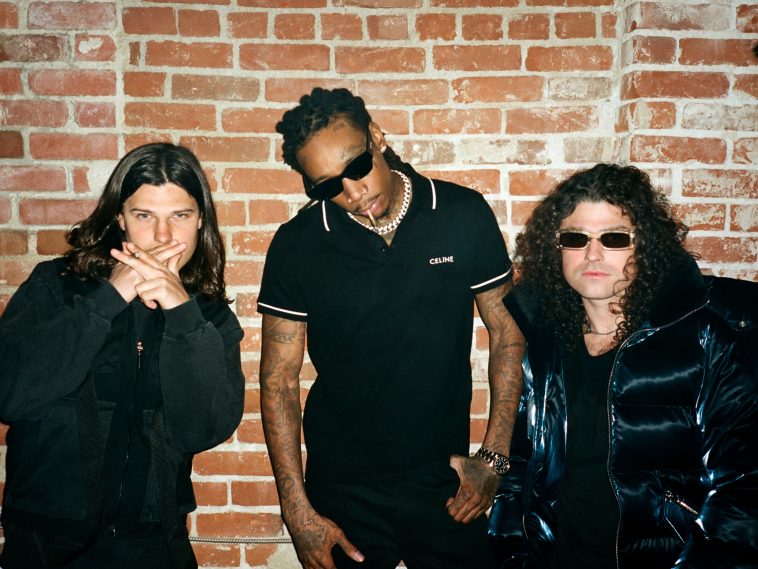Promotional photo for "Sh Sh Sh (Hit That)" which sees DVBBS posing with Wiz Khalifa against a bright wall, with the rapper in the middle. All three are wearing black, with Wiz Khalifa and one-half of DVBBS donning shades.