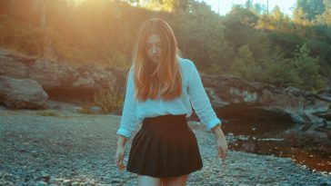Promotional photo for "Alleyways" which sees Faith Marie with ginger mid-length hair falling over a white long-sleeve shirt that is stuck into a black ra ra skirt. She's walking on a rocky beach with trees and greenery in the background.