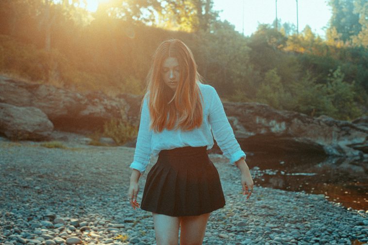 Promotional photo for "Alleyways" which sees Faith Marie with ginger mid-length hair falling over a white long-sleeve shirt that is stuck into a black ra ra skirt. She's walking on a rocky beach with trees and greenery in the background.