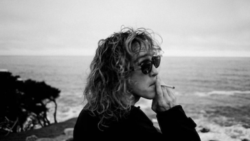 Promotional photo for "Free Love" that is a black and white side-shot of Felly wearing sunglasses and a jumper, smoking a cigarette as he watches the sea and the surf.