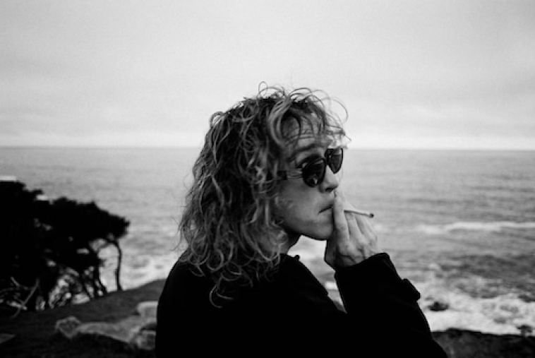 Promotional photo for "Free Love" that is a black and white side-shot of Felly wearing sunglasses and a jumper, smoking a cigarette as he watches the sea and the surf.