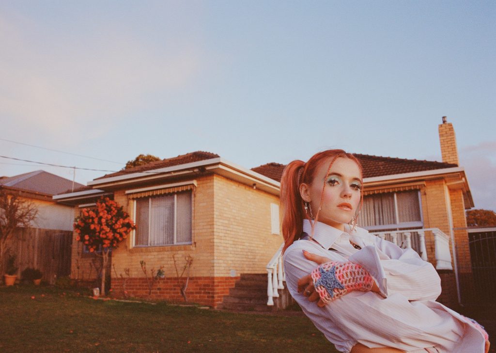 Promotional photo for "This Is Love" EP which sees Hallie posing to the right of the image, in front of a bungalow brick building. Her orange hair is tied up in ponytails and she is wearing a white jacket.