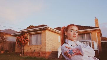 Promotional photo for "This Is Love" EP which sees Hallie posing to the right of the image, in front of a bungalow brick building. Her orange hair is tied up in ponytails and she is wearing a white jacket.