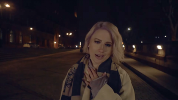 Screenshot from the official music video for "Could Be The One" which sees Jill Marie Cooper walking down the street at night with a cream coat on and a checkered scarf. She has her left hand on her heart as she sings at the camera.