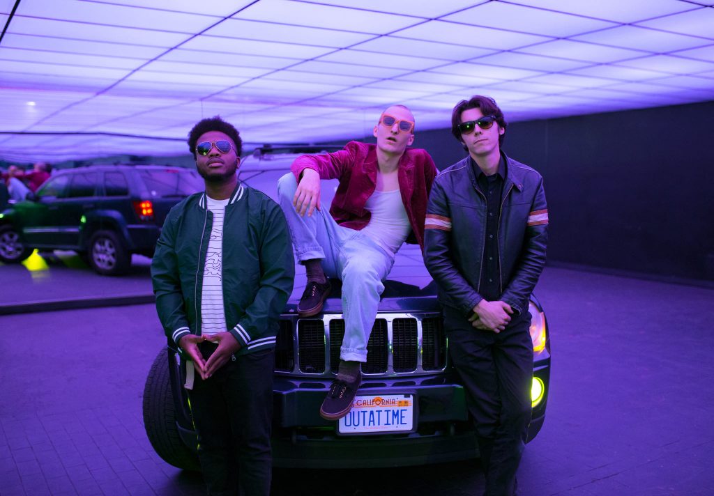 Promotional photo for "Medicine" which sees Nxtime leaning against the front of what looks like a Range Rover with Ray Rubio on top of the bonnet wearing a red jacket, a white vest, white-blue jeans and black combat boots, he has one arm resting on a raised knee as he leans backwards. Nate Christensen is on the right, wearing a black leather jacket with black jeans and a black top, and Imhotep Williams is on the left wearing a white t-shirt under a bright green jacket and black jeans. All three are wearing dark shades.