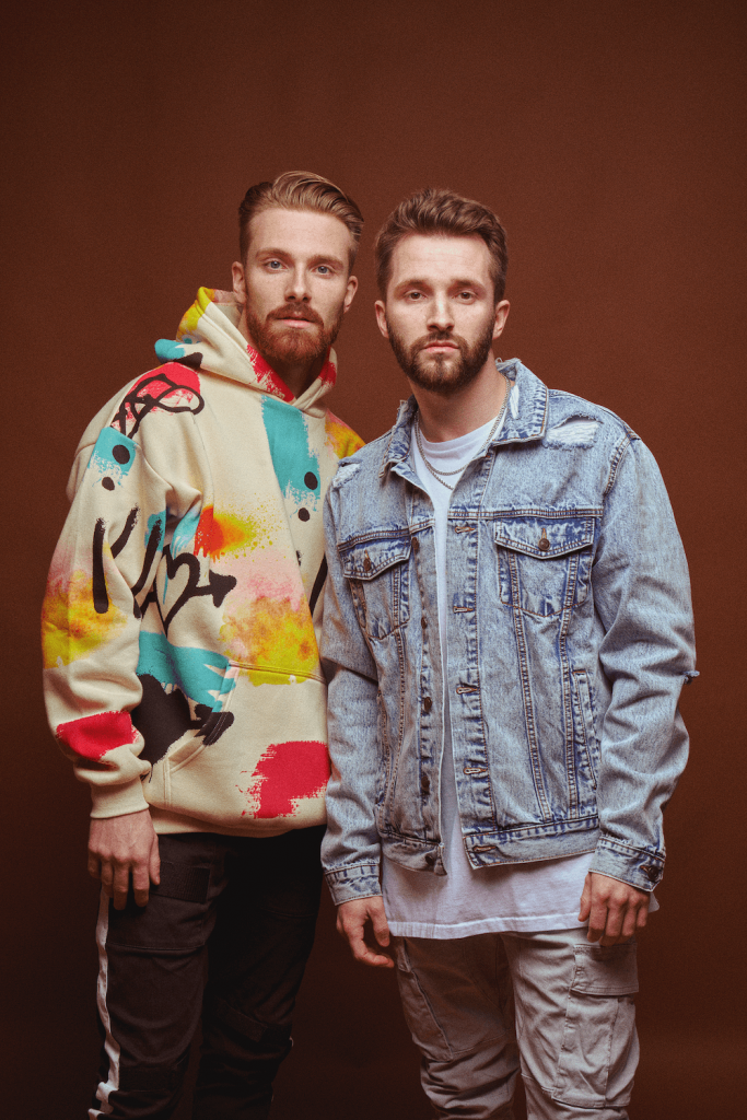 Promotional photo for "holiday." which sees RAGS AND RICHES standing in front of an orange-brown background. Peyton is slightly taller than his brother with a ginger quiff and a beard, and he is wearing a cream hoodie which has blue, red, and yellow squares over it in some sort of pattern. Tanner has brown hair in a similar style, a beard, and he is wearing a white t-shirt under a blue denim jacket.