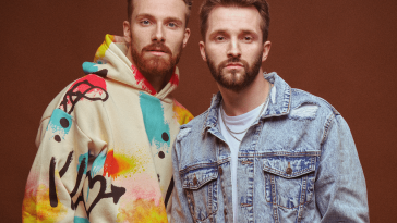 Promotional photo for "holiday." which sees RAGS AND RICHES standing in front of an orange-brown background. Peyton is slightly taller than his brother with a ginger quiff and a beard, and he is wearing a cream hoodie which has blue, red, and yellow squares over it in some sort of pattern. Tanner has brown hair in a similar style, a beard, and he is wearing a white t-shirt under a blue denim jacket.