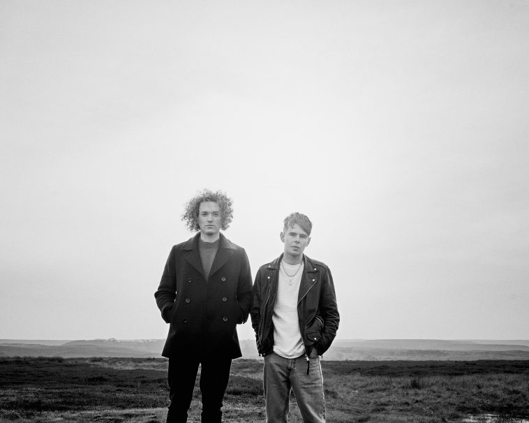 Promotional photo for "Remind Me to Forget You" which is a black and white photo of Seafret standing on a beach, amongst the rock pools, with hands in their pockets, looking at the camera with their black jackets wrapped around them - it's clearly cold.