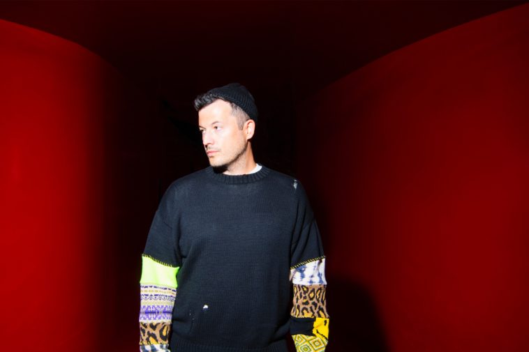 SYML posing in front of a dark red background, wearing a black t-shirt over a sweatshirt that has various different animal prints going down the sleeves, paired with a beanie hat, in promotion of his sophomore album "The Day My Father Died".