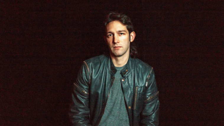 Promotional photo for "Face The Music" which sees Young-Borra posing against a dark backdrop. He has his brown hair styled as a mullet with a quiff fringe. He's wearing a dark green t-shirt under a black leather jacket.
