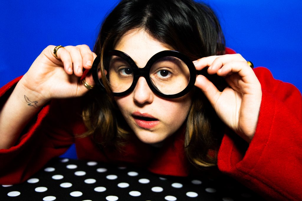 Promotional photo for "Cool Whip" which sees Belot posing at a table with a black with white polka dot table cloth, backgrounded by a blue background, as she leans forward putting on some black-framed circular glasses and she has her tongue just on the inside of her mouth, clearly not believing a word she's being told. She's wearing a red jumper which emphasises her gorgeous wavy brown hair.