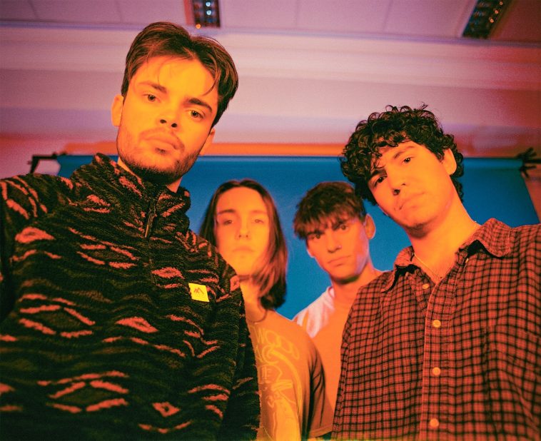 Promotional photo for "Love in the Afternoon" which sees the four-piece band, Blondes, crowded around the camera, looking somewhat down. Will Potter is wearing a hoodie with his dark hair in curtains hairstyle, while the rest of the band are wearing shirts and are slightly back, all with dark hair and different hairstyles, one has shoulder-length hair, one has short curly hair, and the other has a straight simple hair cut.
