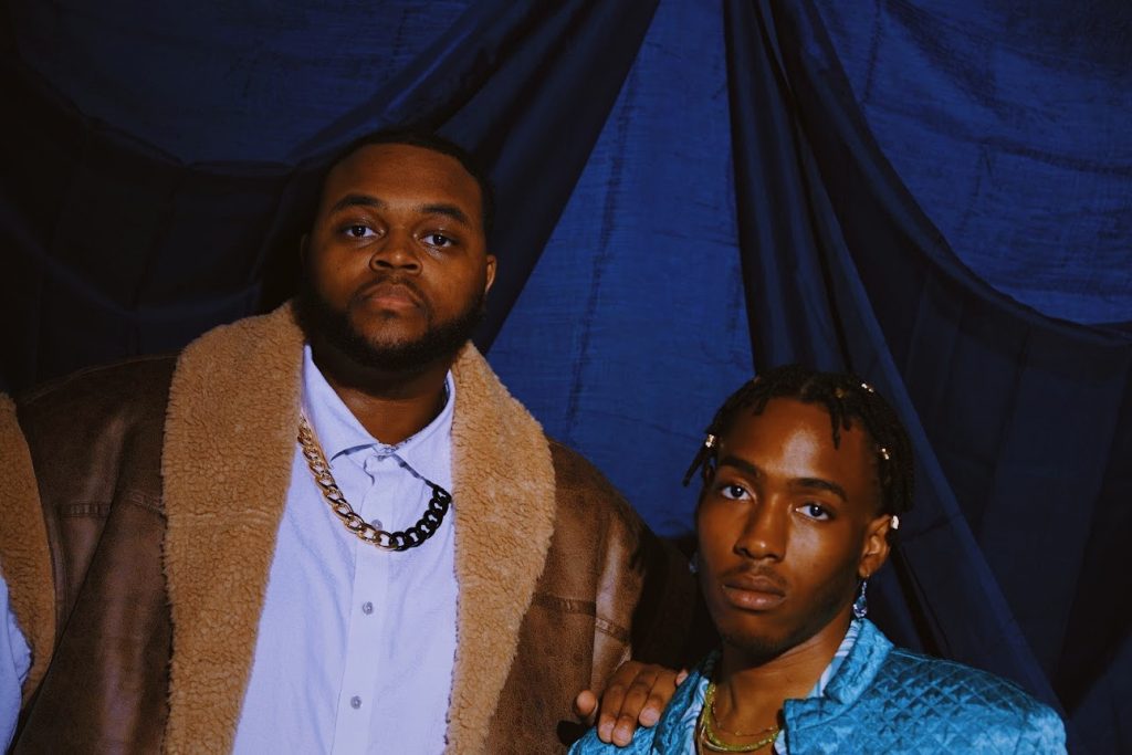 Promotional image for "That Bitch Remix" which sees Cedric St Louis on the right, sitting down, with Fat Westbrook on the left, with his hand on Cedric's shoulder. Cedric is wearing a bright blue shirt, while Fat Westbrook is wearing a white buttoned-up shirt under a gold chain and a wooly brown coat.