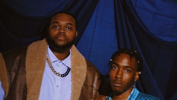 Promotional image for "That Bitch Remix" which sees Cedric St Louis on the right, sitting down, with Fat Westbrook on the left, with his hand on Cedric's shoulder. Cedric is wearing a bright blue shirt, while Fat Westbrook is wearing a white buttoned-up shirt under a gold chain and a wooly brown coat.