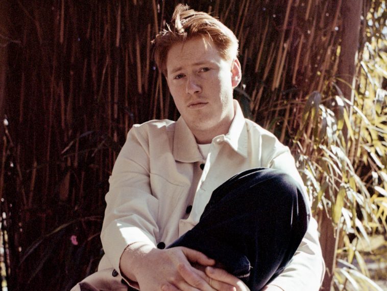 Promotional photo for "Emotional Punching Bag" which sees dePresno sitting down and leaning against a tree trunk with one knee up against his chest while his hands are clasped around his leg. He has ginger styled hair that has been ruffled by the wind into a quiff, while he is wearing a cream light jacket and black jeans.