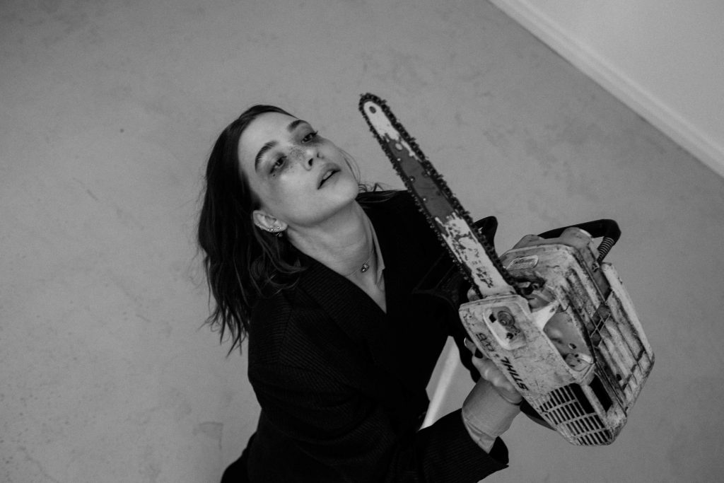 Promotional photo for "Heavy Objects" which is a black and white photo of Dottie Andersson holding up an electrical saw in the air, with her head tilted backwards with her dark hair cascading down her back. She is wearing a black jacket.