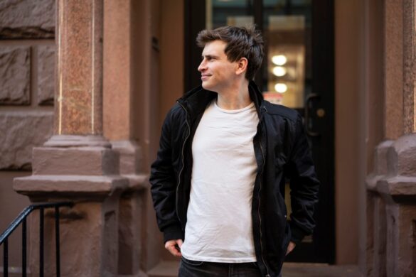 Promotional photo for "Anna" which sees Ilan Bell stepping out of a building, wearing a tight white t-shirt under a black jacket, whilst looking down the street to the left.