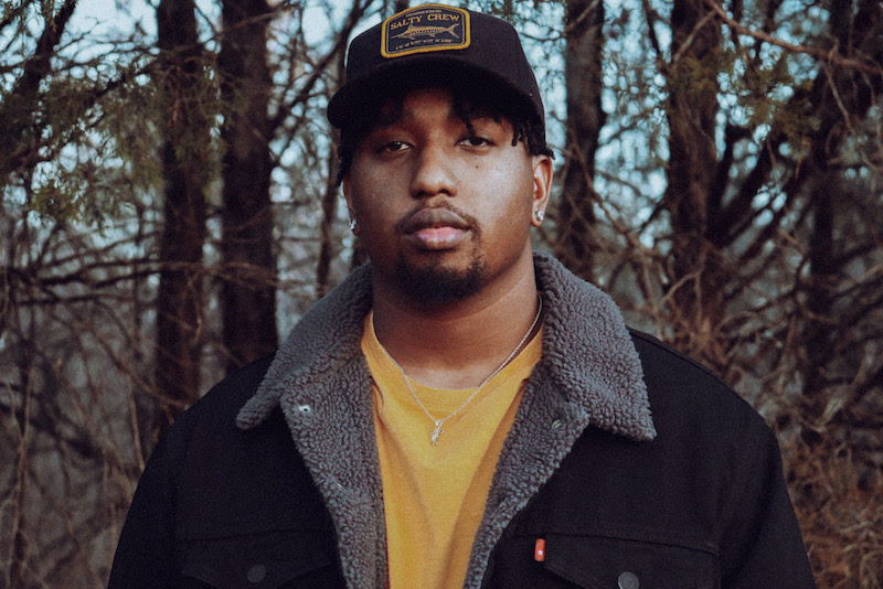 Promotional photo for "Reverse" which sees RVSHVD standing in a forest with a black trucker snapback cap, a sunshine yellow t-shirt underneath a black warm jacket.