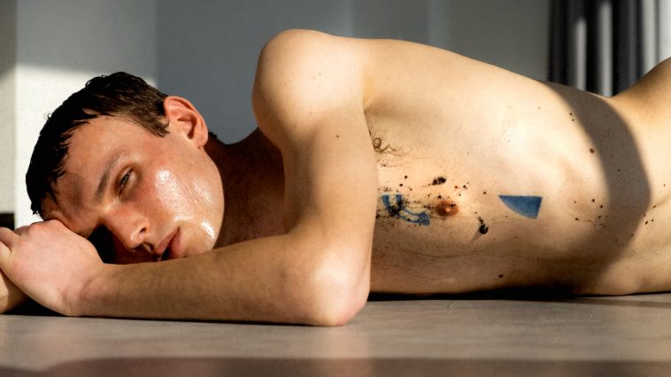 Promotional photo for "My Sting Machines" and "Lots if Love" which sees Scott Wade lying on the floor, face down and shirtless, lying slightly on his side as he looks at the camera. His body is wet and he has dirt trailing down his body, amongst a blue triangle tattoo with a blue quarter-circle tattoo above his nipple. He is resting his head against one of his arms.