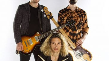 Promotional photo for "CRANIOTØMY" which sees the band SØUNDHOOSE standing as a trio for a photo shoot. Two band members, Brandon Smith and Jeremy Bauer are standing with their guitars over opposite shoulders so the guitar necks cross in the middle of the image. In front of them is Paul Burns who is kneeling down. He has shoulder-length curly blonde hair and he has his drumsticks in his hands and is posing with them in an X presentation.