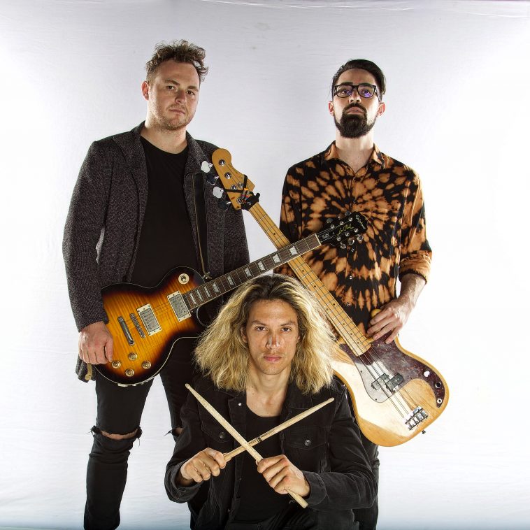 Promotional photo for "CRANIOTØMY" which sees the band SØUNDHOOSE standing as a trio for a photo shoot. Two band members, Brandon Smith and Jeremy Bauer are standing with their guitars over opposite shoulders so the guitar necks cross in the middle of the image. In front of them is Paul Burns who is kneeling down. He has shoulder-length curly blonde hair and he has his drumsticks in his hands and is posing with them in an X presentation.
