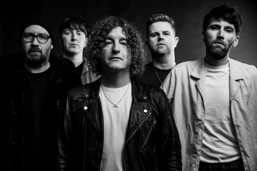 A black-and-white promotional photo for "Lovers Come and Lovers Go" which shows The Pigeon Detectives all lined up with Matt Bowman in the front, whilst two other band members stand shoulder-length apart behind him, and then the other two band members fill the gap between the band members' shoulders, at the back of the photo.