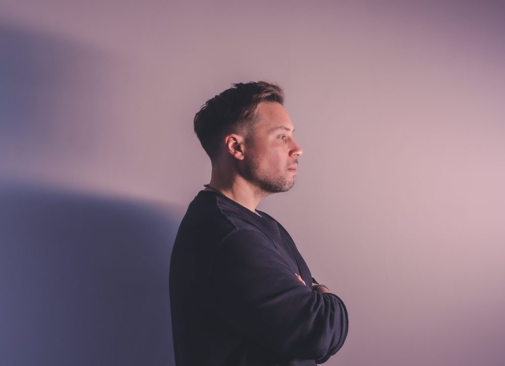 Promotional photo for Balance 31 and "Eteri" which sees Tim Green wearing a navy sweater and facing to the right with his arms crossed, while the photo studio background is a lilac purple.