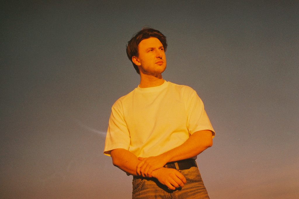 Promotional photo for "Carousel" which sees Tobias Arbo looking off to the side with his arms held out in front of him. He is wearing a white t-shirt and grey jeans, with an ombre sunset-like background.