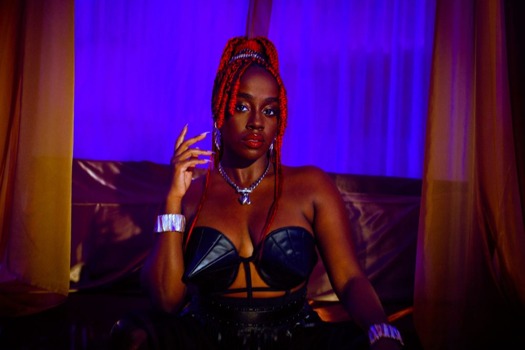 Promotional photo for "Cocoa Butter" which sees AIZA with red hair, wearing a black cone bra, sitting on a brown couch with a neon purple haze lighting going on in the background.