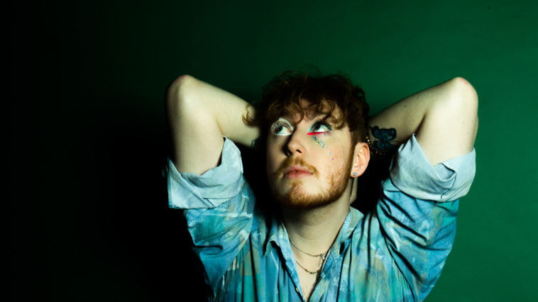 Promotional photo for "To Miss You" which sees Blair Davie posing against a green background with his arms behind their head, wearing a blue denim shirt with the sleeves rolled up. He has dark ginger curly hair, a short beard, and colourful eyeliner.