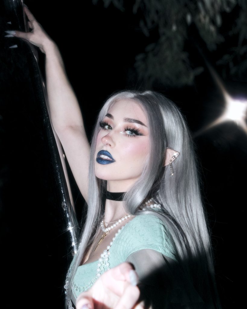 Promotional photo for "Watch Me" which sees bludnymph wearing a light blue bra top, and silver hair, holding onto a metal pole as she looks at the camera.