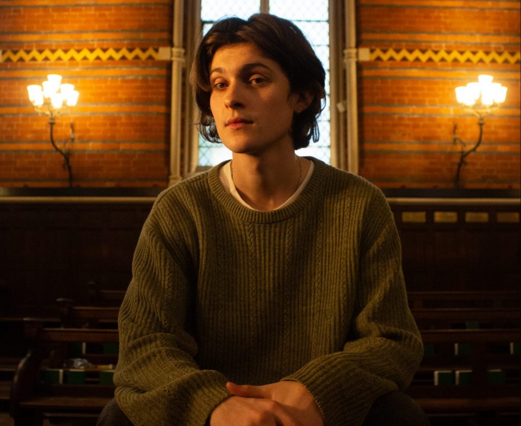 Promotional photo for "The Rites" which sees bryden posing in front of a window at a chapel, sitting down on a pew. He is wearing a green sweater.