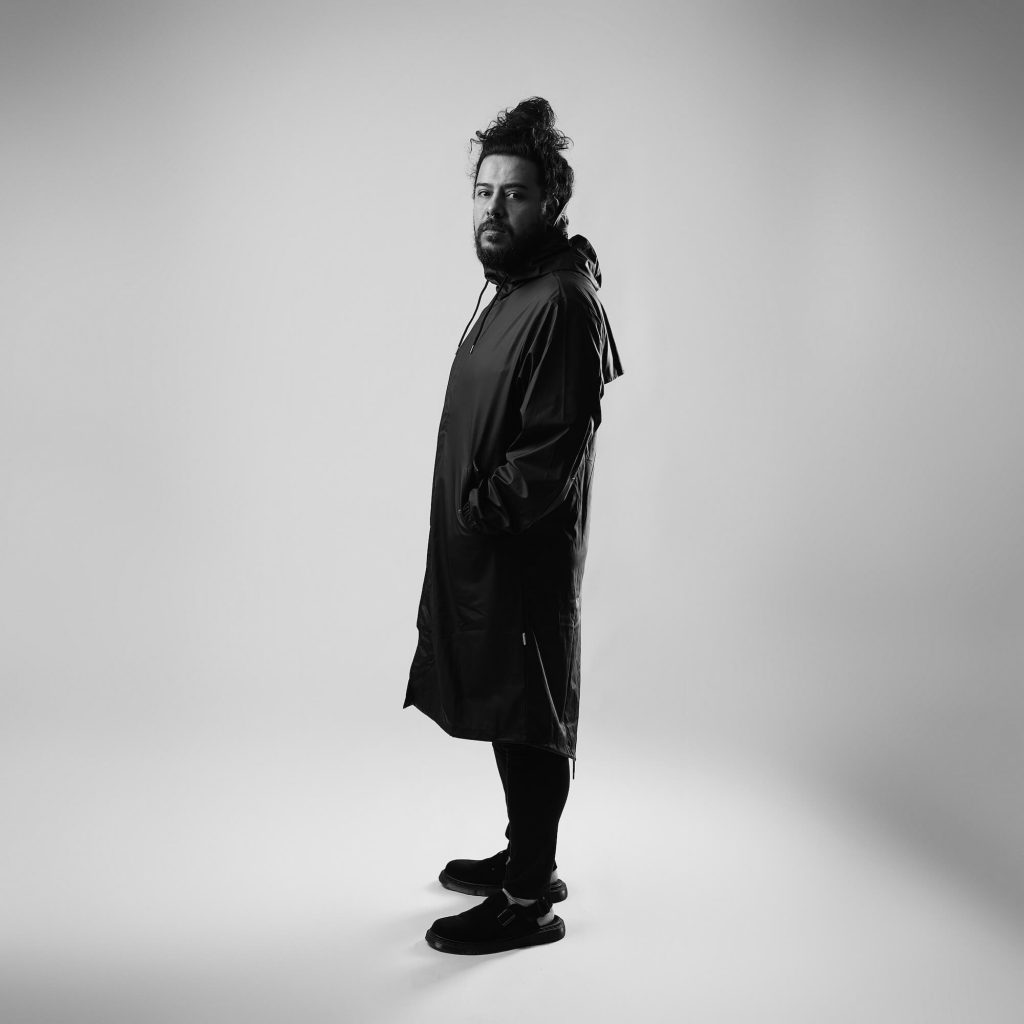 Promotional photo for "I'm Glad I Found U Pt.1" which sees GANZ standing to the left, decked out in a black mac trench coat with black trousers and black trainers, with a white background. He is slightly looking towards the camera.