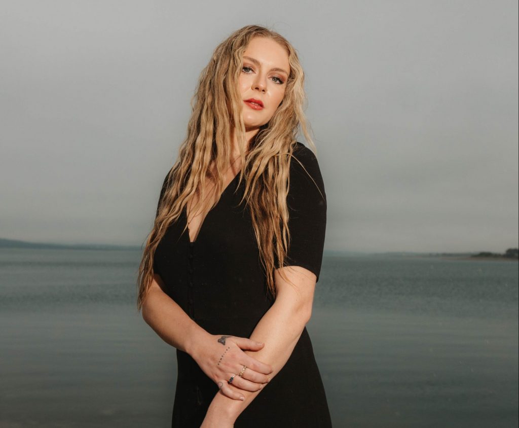 Promotional photo for "how we used to be" which sees Emily Morsen posing with the sea behind her. She's wearing a black dress with a big open-cleavage section, while her long wavy we hair is falling past her elbows.